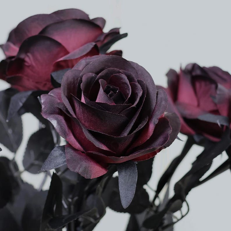 May include: Close-up of artificial roses with deep burgundy petals and dark gray leaves. The roses have a velvety texture and are in full bloom, showcasing their intricate layers. The stems are a dark gray color, and the background is a soft white.