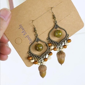 May include: A pair of bronze-toned chandelier earrings featuring a teardrop-shaped frame, a central olive green bead, and a dangling acorn charm. The design includes small brown and cream-colored wooden beads along the bottom edge.