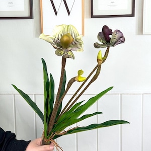May include: A bouquet of artificial orchids featuring two open blooms and several buds. The flowers have a mix of green, purple, and yellow hues, with textured petals and brown stems. The bouquet includes long, green leaves.