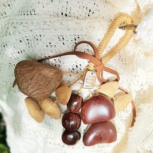 May include: A collection of natural elements, including a large brown nut, various seeds, and polished, reddish-brown stones. A woven, heart-shaped element and a small tag with text are also present. The items are arranged on a white, textured surface.