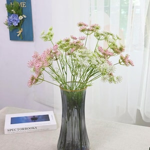 May include: A glass vase with a gold rim holds a bouquet of pink, white, and green flowers. The vase is a smoky gray color and sits on a woven coaster. A book with the text "SPECTORBOOKS" is visible.