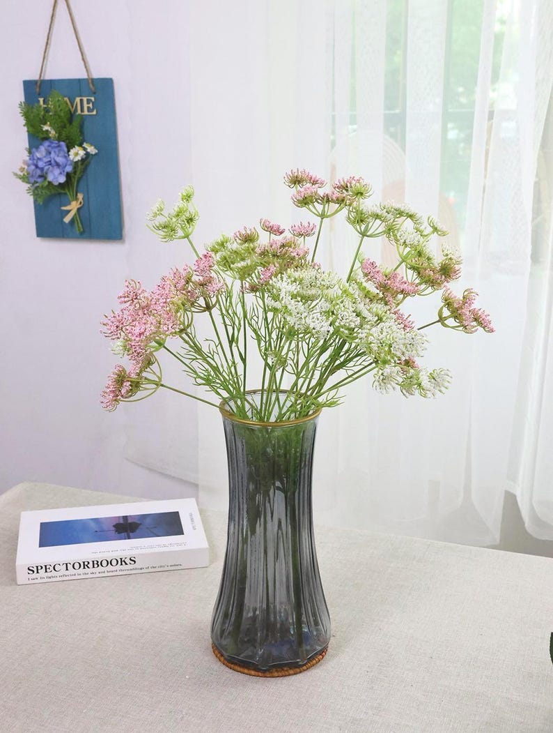 May include: A glass vase with a gold rim holds a bouquet of pink, white, and green flowers. The vase is a smoky gray color and sits on a woven coaster. A book with the text "SPECTORBOOKS" is visible.
