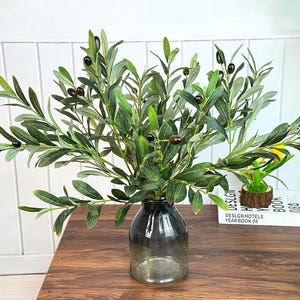 May include: A glass vase displays olive branches with green leaves and dark olives. The vase is on a wooden table, creating a natural display. The arrangement adds a touch of nature to any space. The table is made of wood.
