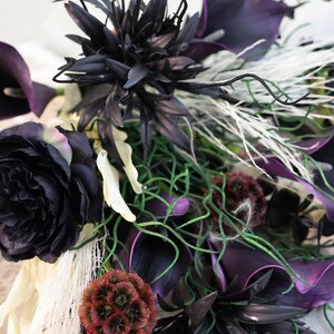 May include: A close-up of a dark floral arrangement featuring black and deep purple flowers, including calla lilies and roses. The bouquet also includes green vines, white feathery accents, and a textured, reddish-brown spherical bloom.