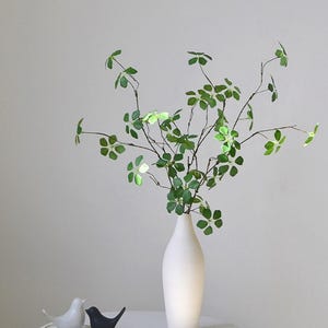 Pu&ograve; includere: Un vaso di ceramica bianco contiene rami verdi e frondosi. Due piccole statuette di uccelli, una bianca e una nera, sono su un piccolo vassoio bianco. La composizione &egrave; su un tavolo bianco su sfondo neutro.