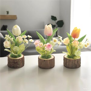 Peut inclure: Trois compositions florales artificielles avec des tulipes, des roses et des marguerites. Chaque arrangement est placé dans une petite base ronde en bois. Les tulipes sont blanches, roses et jaunes. Les roses sont roses et jaunes. Les marguerites sont blanches.