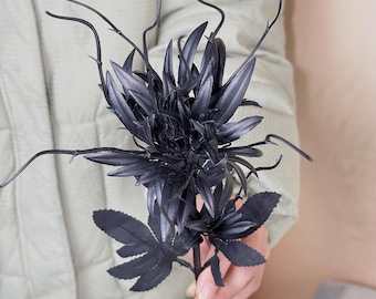 May include: A black artificial flower with multiple spiky leaves and thin, curved stems. The flower is held in a person's hand, and the background is a light beige color. The flower's stem is long and thin.