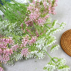 May include: Close-up of artificial flower stems with pink and white blossoms. The flowers have green stems and leaves. The arrangement is on a light gray surface, with a woven basket in the background. The flowers are likely for home decor.