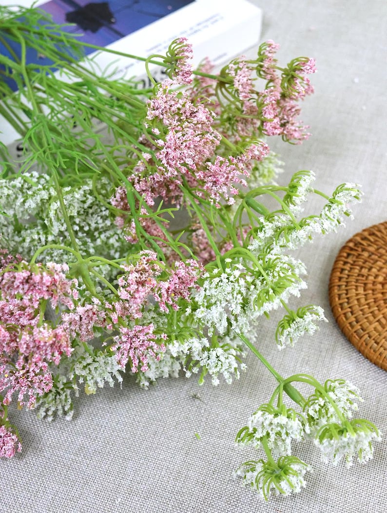 May include: Close-up of artificial flower stems with pink and white blossoms. The flowers have green stems and leaves. The arrangement is on a light gray surface, with a woven basket in the background. The flowers are likely for home decor.