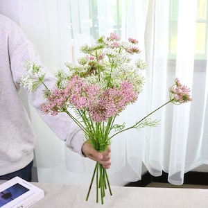 May include: A bouquet of artificial flowers featuring a mix of pink and white blossoms with green stems. The flowers are held together, creating a visually appealing arrangement. The background includes a white curtain and a table.