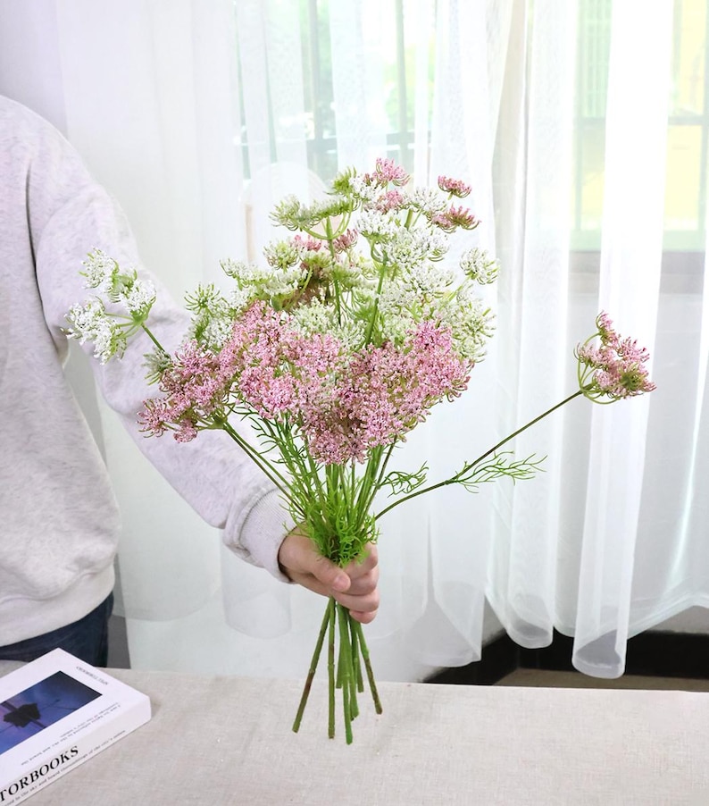 May include: A bouquet of artificial flowers featuring a mix of pink and white blossoms with green stems. The flowers are held together, creating a visually appealing arrangement. The background includes a white curtain and a table.