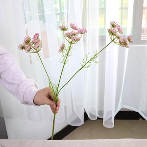 May include: A sprig of artificial flowers with small, pink, clustered blooms. The stems are green, and the flowers are held by a person's hand. The background features a sheer white curtain.