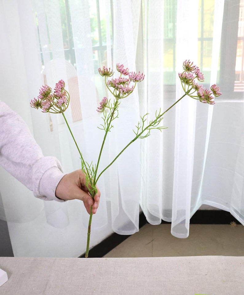 May include: A sprig of artificial flowers with small, pink, clustered blooms. The stems are green, and the flowers are held by a person's hand. The background features a sheer white curtain.