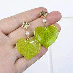 May include: A pair of drop earrings featuring heart-shaped lime green leaves encased in a clear resin finish. Each earring includes a small white pearl bead and a textured gold-tone square stud component at the top, connected by gold-tone metal links.