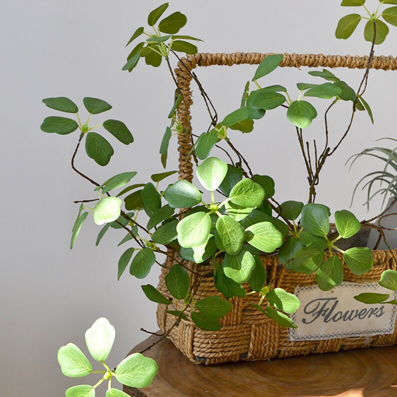 Pu&ograve; includere: Un cesto intrecciato pieno di piante artificiali con foglie verdi. Il cesto ha una forma rettangolare e un manico in corda ritorta. Una piccola etichetta con la parola "Flowers" &egrave; attaccata al cesto.