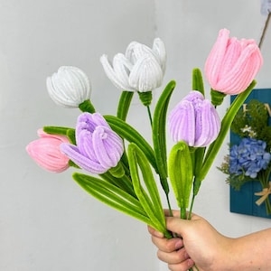 May include: A bouquet of handmade tulip flowers in shades of white, pink, and purple. The flowers have a textured, fuzzy appearance, with green stems and leaves. The bouquet is held in a hand.