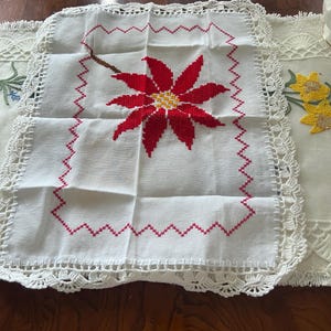 May include: White crocheted square table runner with a red poinsettia embroidered in the center. The runner has a pink zig-zag border.
