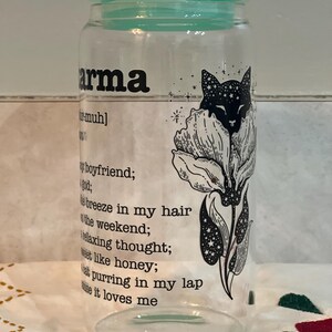 May include: A clear glass jar with a teal lid and a black and white illustration of a cat in a flower. The jar has the word "karma" printed on it and a definition of the word below the illustration.