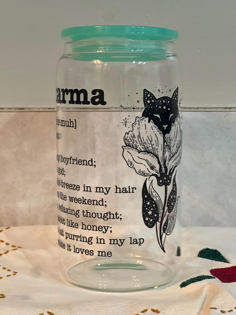 May include: A clear glass jar with a teal lid and a black and white illustration of a cat in a flower. The jar has the word "karma" printed on it and a definition of the word below the illustration.