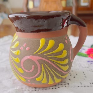 May include: A small, handcrafted ceramic pitcher with a brown rim and handle. The body is tan with yellow and pink floral designs. The pitcher has a rounded shape and is likely used for serving beverages.