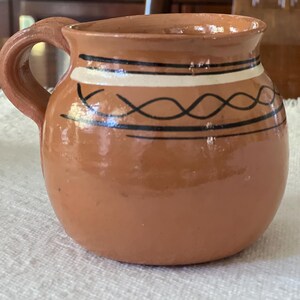 May include: A small, round, terracotta-colored mug with a handle. The mug is decorated with black and white horizontal bands and a black wavy design. The mug is sitting on a white, textured surface.