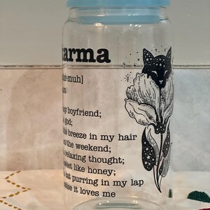 May include: A clear glass jar with a blue lid and a black and white illustration of a cat with stars and a flower. The jar has the word "karma" printed on it with a definition below.