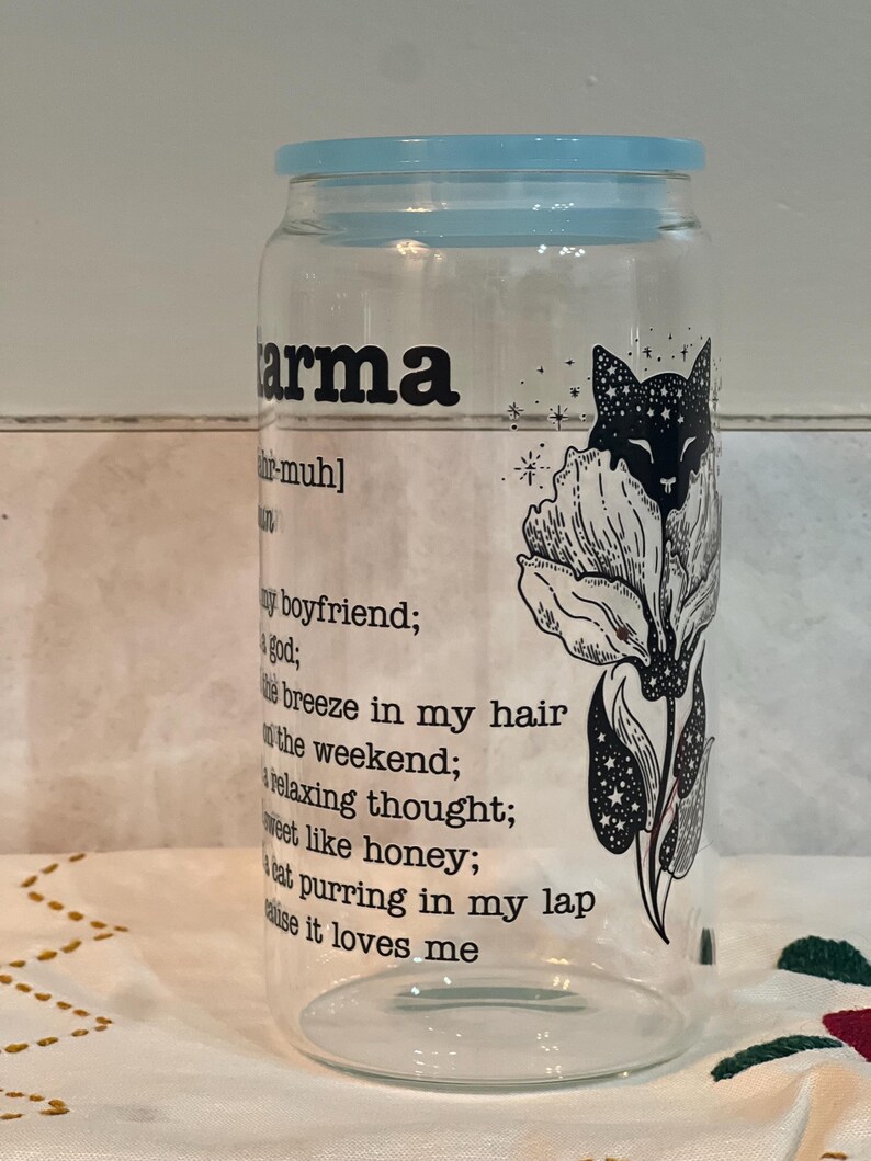 May include: A clear glass jar with a blue lid and a black and white illustration of a cat with stars and a flower. The jar has the word "karma" printed on it with a definition below.