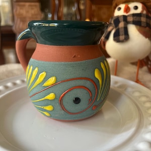 May include: A small, hand-painted ceramic mug with a handle. The mug has a teal base with yellow and orange floral designs, a terracotta band, and a dark green rim. It sits on a white plate.