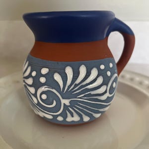 Op de afbeelding: Een kleine terracotta kan met een donkerblauwe rand en handvat. Het onderste gedeelte is lichtblauw geschilderd met een handgeschilderd wit bloemen- en krulpatroon. De kan heeft een ronde vorm met één handvat en een brede opening.