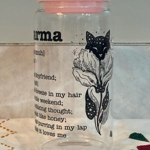 May include: A clear glass jar with a pink lid and a black and white illustration of a cat in a flower. The text on the jar reads "arma [muh] boyfriend; breeze in my hair the weekend; relaxing thought; sweet like honey; cat purring in my lap cause it loves me".