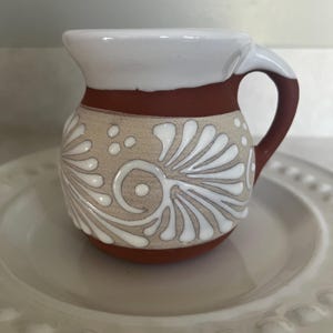 Puede incluir: Una jarra pequeña de terracota con borde y asa esmaltados en blanco. El cuerpo presenta una banda beige decorada con motivos florales en espiral y puntos pintados en blanco. La jarra tiene una forma redondeada y un asa en el lado derecho.