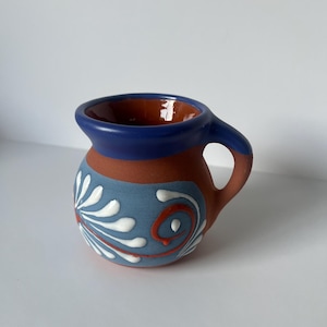 May include: A small terracotta mug with a rounded body and a handle. The rim is painted in a solid dark blue. The exterior features a light blue band decorated with white painted floral and swirl patterns, contrasting with the natural clay color.