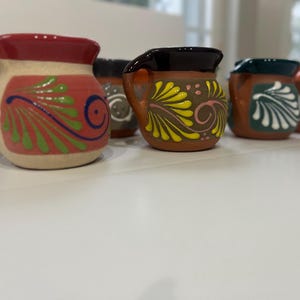 May include: A selection of small, hand-painted ceramic mugs. The mugs display vibrant designs in red, green, yellow, and white against a brown and beige background. Each mug has a handle and a unique decorative pattern, showcasing a traditional aesthetic.