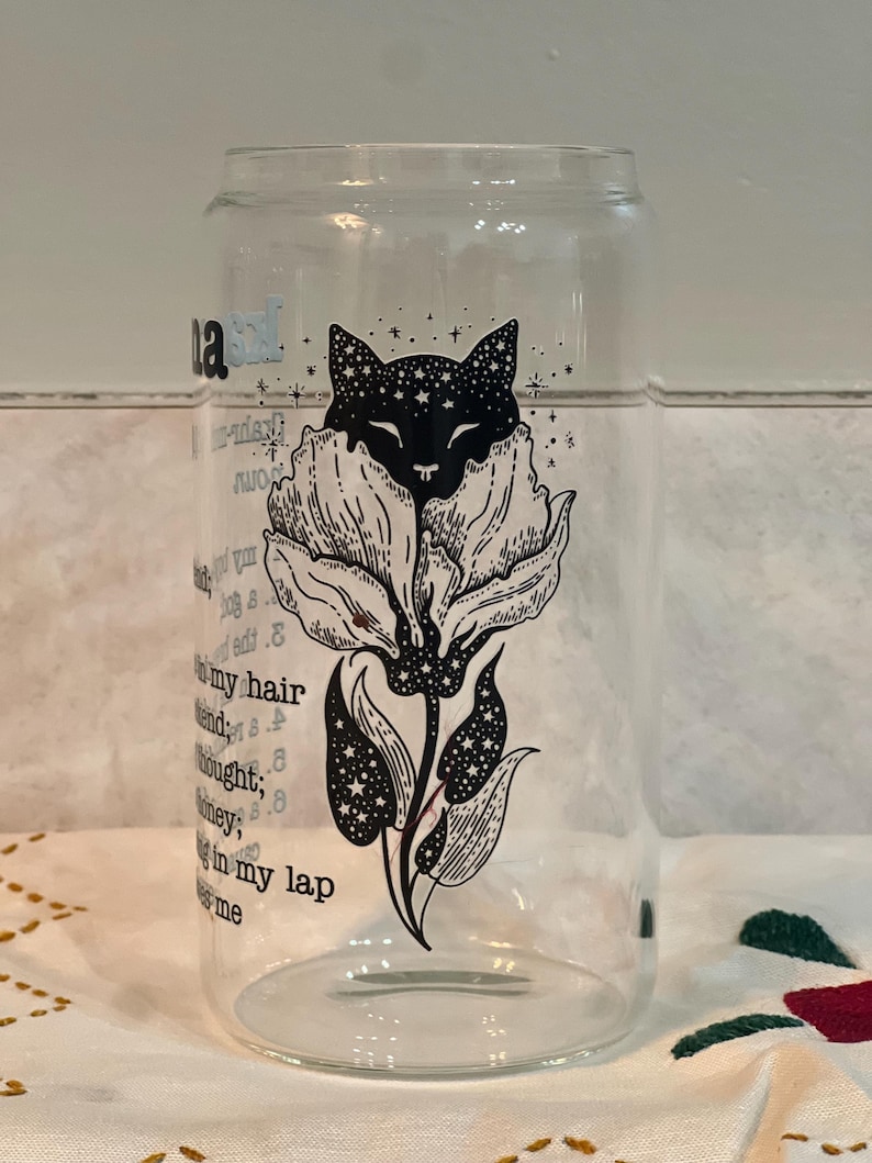 May include: A clear glass cup with a black and white illustration of a cat with stars in its fur, inside a flower. The text "Sleep in my hair, friend; thought, honey; Sleep in my lap, me" is printed on the cup.
