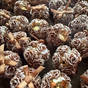 May include: A collection of small, decorative rose-shaped items in shades of brown and cream. Some are adorned with faux pearls, gold-colored ribbons, and heart-shaped or gem-like embellishments. The items appear to be handmade and are arranged closely together.