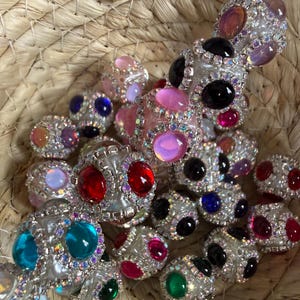 May include: A collection of decorative skewers with colorful, oval-shaped gemstones. The gems are set in silver-colored metal and surrounded by smaller, clear crystals. The skewers are displayed in a woven basket.