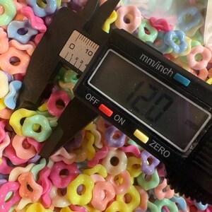 Puede incluir: Primer plano de una colecci&oacute;n de cuentas en forma de anillo de colores pastel. Un calibrador digital negro mide las cuentas, mostrando una medida de 12,0 mm. Las cuentas son de color rosa, amarillo, azul y naranja.