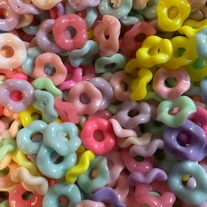 Puede incluir: Primer plano de una colecci&oacute;n de cuentas de pl&aacute;stico de colores en varias formas. Las cuentas son en tonos pastel de rosa, amarillo, azul, verde y morado. Las formas incluyen anillos, flores y dise&ntilde;os ondulados. Ideal para la fabricaci&oacute;n de joyas y manualidades.