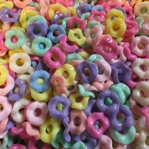 Puede incluir: Primer plano de un mont&oacute;n de cuentas de colores pastel. Las cuentas tienen varias formas, incluyendo anillos, flores y formas retorcidas. Los colores incluyen rosa, amarillo, azul, verde y morado.