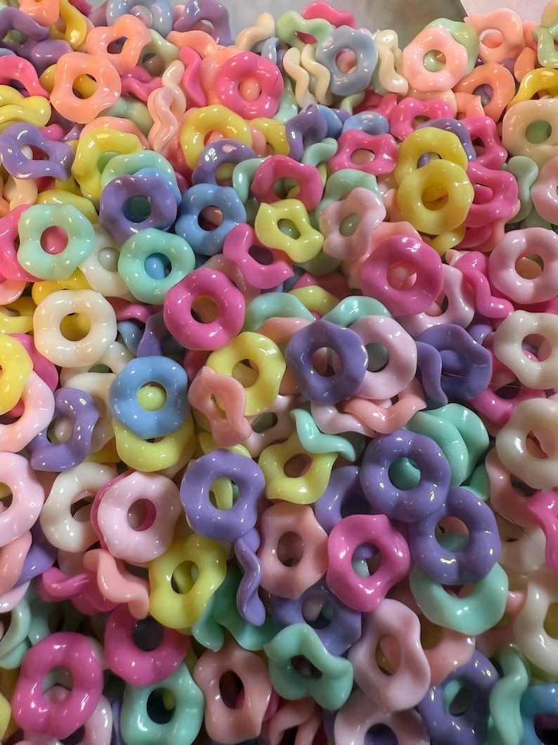 Puede incluir: Primer plano de un mont&oacute;n de cuentas de colores pastel. Las cuentas tienen varias formas, incluyendo anillos, flores y formas retorcidas. Los colores incluyen rosa, amarillo, azul, verde y morado.