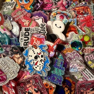 Puede incluir: Una colección de coloridos y brillantes pines y chapas con varios diseños. Los pines presentan personajes de dibujos animados, referencias a la cultura pop y texto como "I'm not rude" y "Sensitive". El surtido incluye una máscara de Jason Voorhees, una lata de Coca-Cola y un personaje de dibujos animados con cabello naranja.