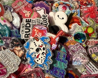 May include: A selection of vibrant, glitter-covered pins and badges with diverse designs. The pins showcase cartoon characters, pop culture themes, and text such as "I'm not rude" and "Sensitive". The collection includes a Jason Voorhees mask, a Coca-Cola can, and a cartoon character with orange hair.