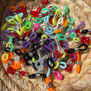 May include: A collection of colorful eyeglass retainers in various shades, including red, orange, yellow, green, blue, pink, purple, and black. Each retainer features a loop design with a silver metal coil. The retainers are scattered in a woven basket.