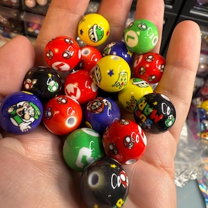 Op de afbeelding: Een verzameling kleurrijke, ronde kralen met verschillende cartoonkarakterontwerpen. De kralen zijn voornamelijk rood, groen, geel, blauw en zwart, met wit en andere accentkleuren. Elke kraal heeft een gat om te rijgen.