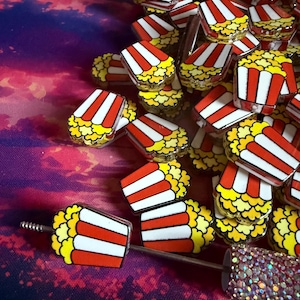 May include: A collection of popcorn-themed accessories. The items feature a red, white, and yellow popcorn design. One item is a pen with a popcorn design and a sparkly, iridescent handle. The other items appear to be decorative pins or charms.