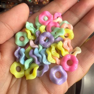 Puede incluir: Un pu&ntilde;ado de cuentas de resina de colores pastel en varias formas, incluyendo rosquillas y espirales. Las cuentas son en tonos rosa, amarillo, verde y morado. Estas cuentas son ideales para la fabricaci&oacute;n de joyas y proyectos de artesan&iacute;a.