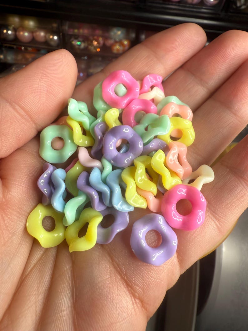 Puede incluir: Un pu&ntilde;ado de cuentas de resina de colores pastel en varias formas, incluyendo rosquillas y espirales. Las cuentas son en tonos rosa, amarillo, verde y morado. Estas cuentas son ideales para la fabricaci&oacute;n de joyas y proyectos de artesan&iacute;a.