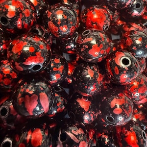 Pode incluir: Um close-up de contas redondas e brilhantes, pretas com salpicos vermelhos vibrantes. Cada conta tem um furo para enfiar. Adequadas para a criação de joias e projetos de artesanato.