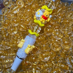 Puede incluir: Un bolígrafo decorativo con un barril blanco y un tema de Winnie the Pooh. El bolígrafo presenta una figura amarilla de Winnie the Pooh, una flor blanca y una sección brillante y con purpurina. El bolígrafo está rodeado de muchas cuentas esféricas pequeñas, claras y amarillas.