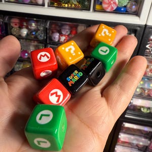 以下が含まれることがあります： スーパーマリオブラザーズのキャラクターとシンボルが描かれた、カラフルなサイコロ7個のセット。サイコロは緑、赤、黒、黄、白です。サイコロは手に持たれています。